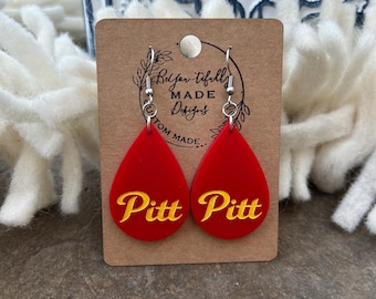 May include: Red teardrop earrings with the word "Pitt" in yellow script. The earrings are displayed on a brown card with the text "Bel for tifully MADE Designs". The earrings have silver-colored hooks.