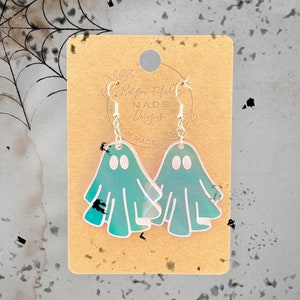 May include: A pair of iridescent green ghost-shaped earrings with silver hooks. The earrings are on a brown card with the text "Blythe Tidbits Made Designs from Made."