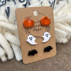 May include: A set of Halloween-themed earrings featuring a pair of orange pumpkins, a pair of white ghosts, and a pair of black bats. The earrings are displayed on a brown card with the text "Custom Made Designs".