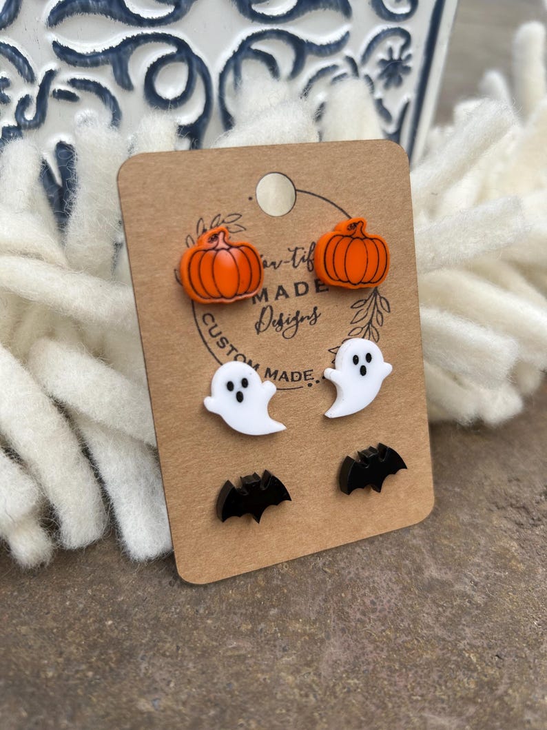 May include: A set of Halloween-themed earrings featuring a pair of orange pumpkins, a pair of white ghosts, and a pair of black bats. The earrings are displayed on a brown card with the text "Custom Made Designs".