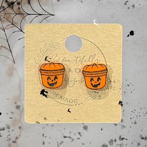 May include: A pair of orange pumpkin-shaped earrings with black jack-o'-lantern faces. The earrings are on a brown card with the text "Red-on-tiffaly AD Designs OM MADE."