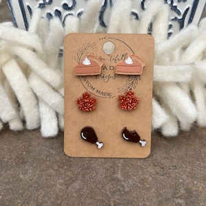 May include: A set of six fall-themed earrings on a brown card. The earrings are made of wood and feature a pumpkin pie, a turkey leg, and a maple leaf. The card has the text "Autumn Tiffani Designs Custom Made."