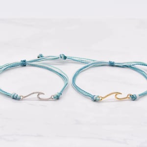 Pode incluir: Duas pulseiras de amizade com um amuleto de onda. Uma pulseira é azul e branca com um amuleto prateado, a outra é azul e branca com um amuleto dourado.