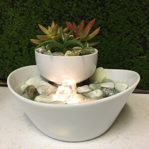 May include: A white ceramic tabletop fountain with a small succulent plant in a white ceramic pot on top. The fountain has a small waterfall and a light that illuminates the water.