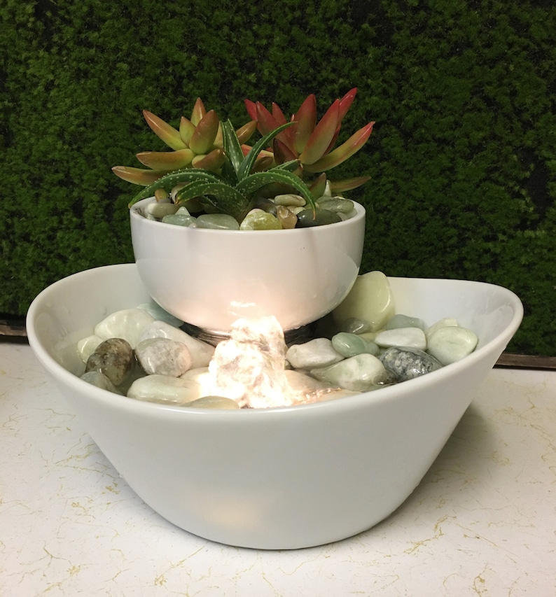 May include: A white ceramic tabletop fountain with a small succulent plant in a white ceramic pot on top. The fountain has a small waterfall and a light that illuminates the water.
