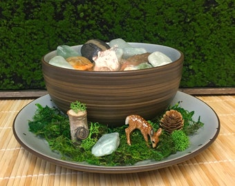 May include: A brown ceramic bowl filled with green and brown stones, surrounded by moss and a small deer figurine. The bowl is sitting on a gray saucer.