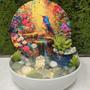 May include: A white bowl fountain featuring a circular art panel with a colorful hummingbird, flowers, and a waterfall. The base contains smooth green and white river stones, a small blue planter with a succulent, and two small green plants.