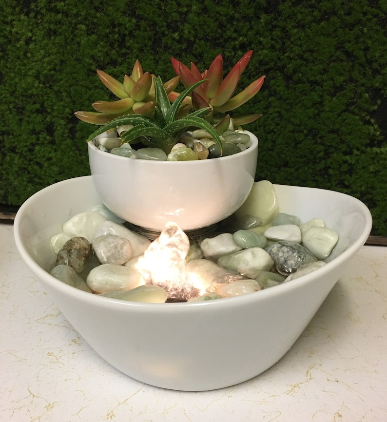 May include: A white ceramic tabletop fountain with a small bowl on top, containing succulents. The bottom bowl is filled with smooth, light-coloured stones and water. The fountain has a small, illuminated water feature.
