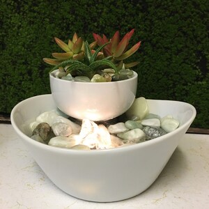 May include: A white ceramic tabletop fountain with a small succulent plant in a white ceramic pot. The fountain has a clear plastic waterfall and white stones in the bottom bowl.
