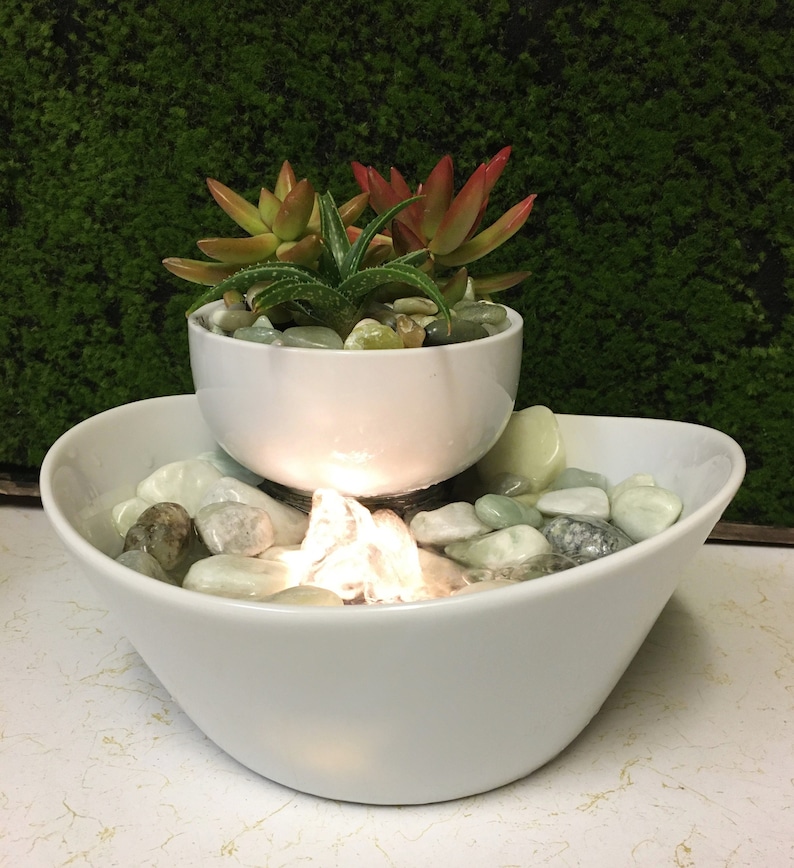 May include: A white ceramic tabletop fountain with a small succulent plant in a white ceramic pot. The fountain has a clear plastic waterfall and white stones in the bottom bowl.