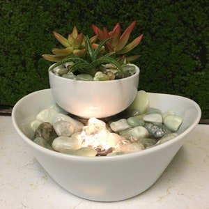May include: A white ceramic tabletop fountain with a smaller bowl on top, containing succulents. The bottom bowl has smooth, light green stones and a glowing light in the centre.