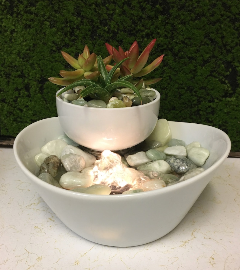 May include: A white ceramic tabletop fountain with a smaller bowl on top, containing succulents. The bottom bowl has smooth, light green stones and a glowing light in the centre.