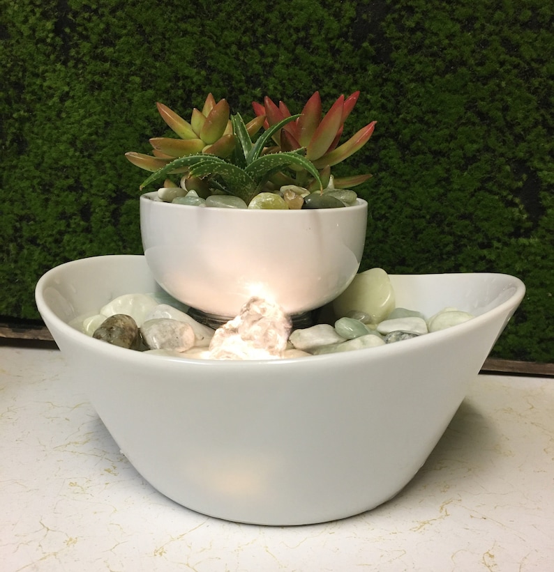 May include: A white ceramic tabletop fountain with a small bowl on top containing succulents and a larger bowl below with smooth white stones and water. The fountain has a light that illuminates the water.