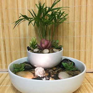 May include: A two-tiered white ceramic fountain with small plants and succulents. The top tier holds a small palm-like plant and a succulent. The bottom tier has water, pebbles, and more succulents. The background is a bamboo mat.