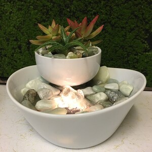 May include: A white ceramic tabletop fountain with a smaller bowl on top, holding succulents. The fountain has a light and a water pump that creates a small waterfall effect. The bottom bowl is filled with smooth, light-colored stones.