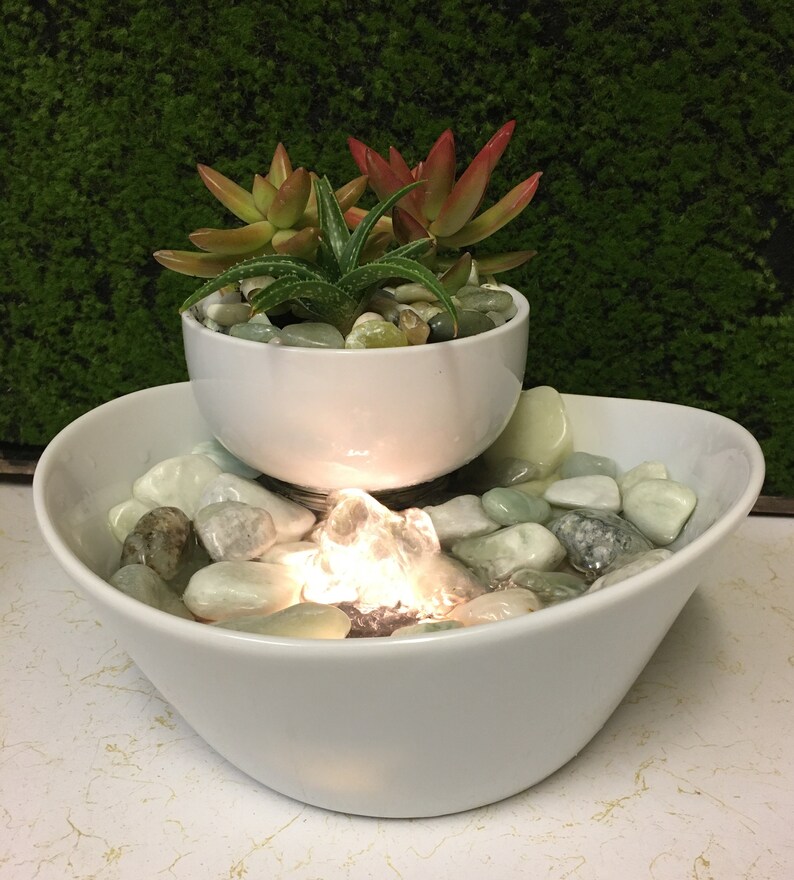 May include: A white ceramic tabletop fountain with a smaller bowl on top, holding succulents. The fountain has a light and a water pump that creates a small waterfall effect. The bottom bowl is filled with smooth, light-colored stones.