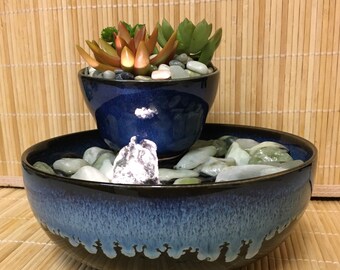 May include: A blue ceramic tabletop fountain with a smaller blue ceramic pot on top. The pot is filled with green and brown succulents and small white stones. The fountain base is filled with water and green stones.