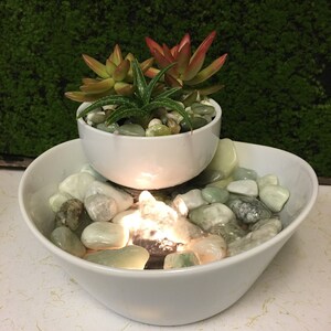 May include: A white ceramic tabletop fountain with a smaller bowl on top, containing succulents. The bottom bowl has a water feature with a light and green stones.