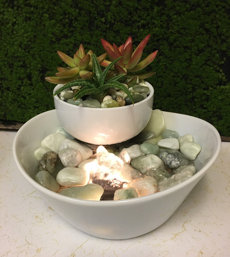 May include: A white ceramic tabletop fountain with a smaller bowl on top, containing succulents. The bottom bowl has a water feature with a light and green stones.