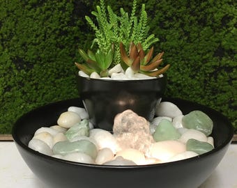 May include: A black ceramic bowl with a smaller black pot inside, filled with succulents and white pebbles. The larger bowl contains white and green stones, with a glowing crystal in the center. The background is a green moss wall.