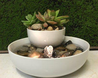 May include: A white ceramic tabletop fountain with a small succulent plant in a white pot on top. The fountain has a small waterfall and is surrounded by smooth gray stones.