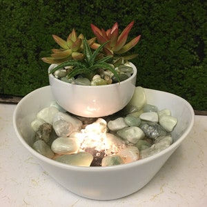 May include: A white ceramic tabletop fountain with two tiers. The top tier features a white ceramic planter with succulents. The bottom tier has a water feature with a light and smooth, light green stones.