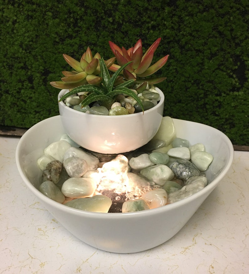 May include: A white ceramic tabletop fountain with two tiers. The top tier features a white ceramic planter with succulents. The bottom tier has a water feature with a light and smooth, light green stones.