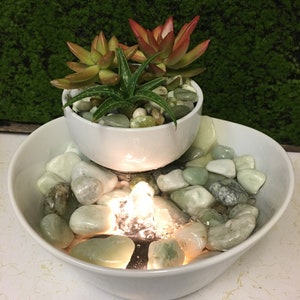 May include: A white ceramic tabletop fountain with a small white planter on top. The planter holds a variety of succulents. The fountain has a small pool of water with smooth, light green stones and a small light that illuminates the water.