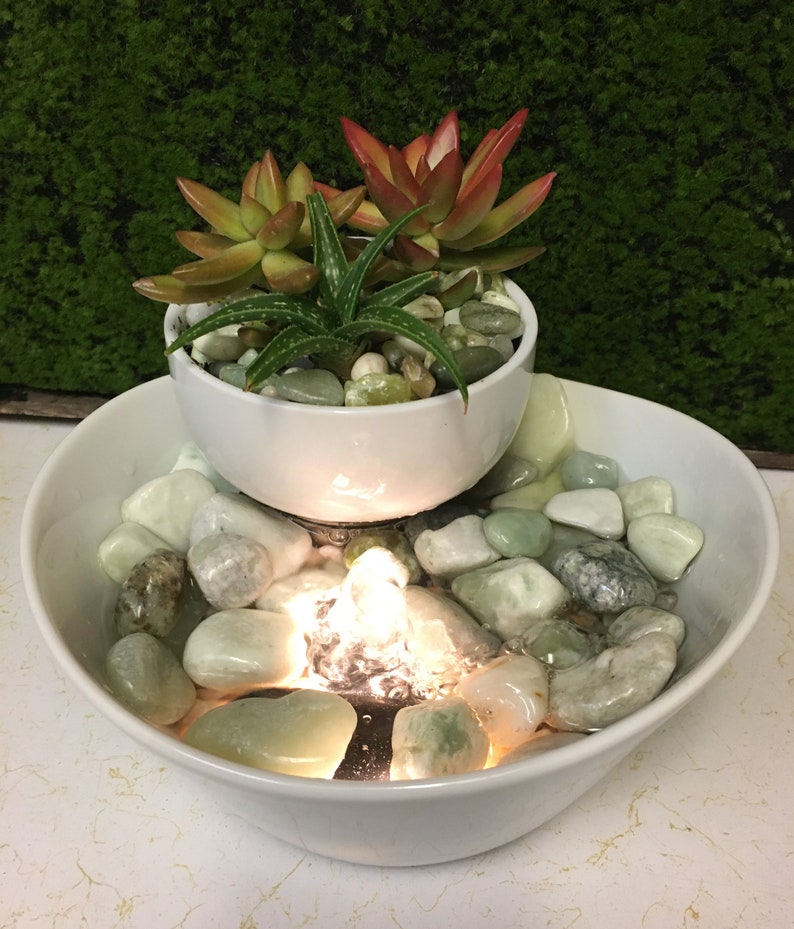 May include: A white ceramic tabletop fountain with a small white planter on top. The planter holds a variety of succulents. The fountain has a small pool of water with smooth, light green stones and a small light that illuminates the water.