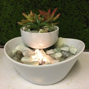 May include: A white ceramic tabletop fountain with a small succulent plant in a white ceramic pot on top. The fountain has a small waterfall and is surrounded by white and green stones.
