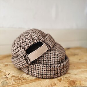 Peut inclure: Deux bonnets à motif à carreaux marron et noir sont empilés. Les bonnets ont un petit bord et une sangle réglable beige. Les bonnets sont en tissu tissé. Ils sont posés sur une surface en bois.