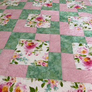 May include: A quilt with a patchwork design featuring alternating squares of pink and green fabric. The green squares have a watercolor floral pattern, while the pink squares have a subtle texture. The quilt is made with a white backing and has a quilted design.