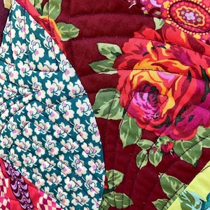 May include: A close-up of a colorful patchwork quilt with a floral pattern. The quilt is made of different fabrics, including a green fabric with white flowers, a red fabric with a floral pattern, and a purple fabric with a paisley pattern.