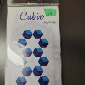 Peut inclure: Un modèle de quilting appelé "Cubix" par Cheryl Phillips, présentant un motif de cubes bleus et violets avec des motifs tourbillonnants. Le motif est imprimé sur du papier blanc et est emballé dans une pochette en plastique transparente.