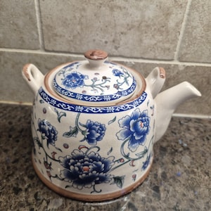 May include: A ceramic teapot with a lid, decorated with blue floral patterns on a white base. It has a brown rim and handle, and a spout. The lid is topped with a small brown knob. The teapot is suitable for serving tea.