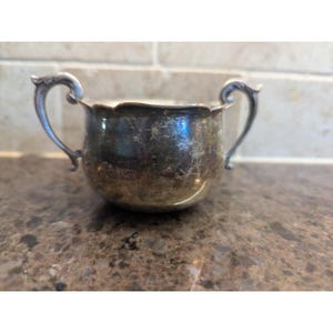 Puede incluir: Un azucarero pequeño de plata con dos asas ornamentadas. El cuenco tiene un borde festoneado y un cuerpo redondeado. La plata tiene un aspecto deslustrado, lo que le da un aspecto vintage. Es probable que el cuenco esté hecho de plata de ley o un metal similar.