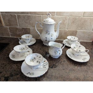 May include: A white porcelain tea set with a floral design. The set includes a teapot, creamer, sugar bowl, and six teacups with saucers. The rims and handles are silver. The floral design is in shades of grey.