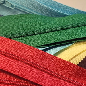 May include: A close-up of a variety of zipper tapes in different colors, including red, green, yellow, blue, and brown. The zippers are arranged in a stack, with the red zipper tape on top.