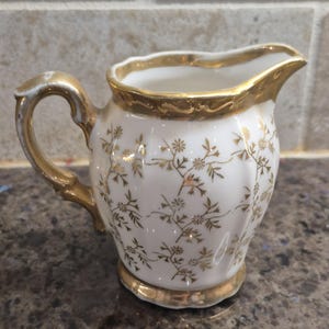May include: A small, white ceramic creamer with gold trim and a gold handle. The creamer is decorated with a delicate gold floral pattern. The rim and base are also trimmed in gold, adding a touch of elegance to the piece.