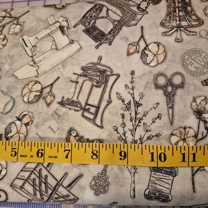 May include: Fabric with a vintage sewing theme. The design features illustrations of sewing machines, scissors, cotton sprigs, and spools of thread in shades of grey and beige. A yellow measuring tape shows inches.