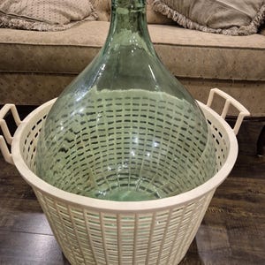 May include: A large, clear, light green glass carboy with a narrow neck and a wide, rounded body, resting inside a round, off-white plastic laundry basket featuring a grid-like pattern and two side handles for carrying and storage.