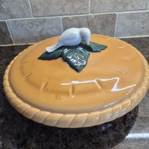 May include: A round ceramic pie dish lid with a glossy tan finish and a braided edge design. The centre features a sculpted white bird perched atop three dark green leaves, serving as a decorative handle for the lid of the baking dish.