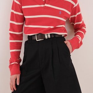 May include: A long-sleeve polo shirt featuring horizontal red and white stripes, a collar with two white buttons, and a small blue embroidered logo on the chest. The shirt is tucked into black pleated shorts secured with a dark brown leather belt.
