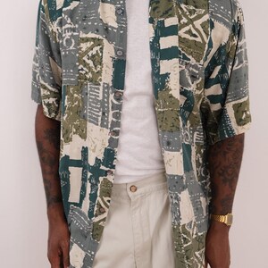 May include: A short-sleeved button-down shirt featuring a multi-colored geometric and abstract block pattern in shades of dark green, olive, cream, and grey. The shirt has a standard collar, a front button placket, and a loose, relaxed fit.