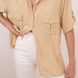 May include: A light beige short-sleeved button-down shirt featuring a classic collar, two front chest pockets with button closures, and a relaxed fit, paired with solid white pants and a gold-toned pendant necklace on a delicate chain.