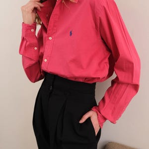 Puede incluir: Una camisa rosa brillante con un pequeño logo azul en el pecho, combinada con pantalones negros de cintura alta. La camisa tiene mangas largas y botones blancos. Los pantalones tienen un frente plisado.