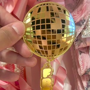 May include: A gold-colored disco ball ornament with a square mirrored surface. The ornament has a gold-colored charm that says "FR". The background includes pink ribbons and floral fabric.