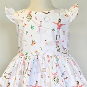 May include: White dress with a festive print featuring ballerinas, nutcrackers, and other Christmas imagery. The dress has a round neckline and short sleeves with ruffles.