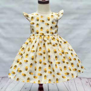 May include: A yellow and white dress with a bee pattern. The dress has short sleeves with ruffles and a full skirt.