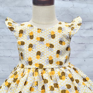 May include: A white dress with a yellow and black bee pattern. The dress has a ruffled neckline and short sleeves. The dress is perfect for a summer party or a day at the park.